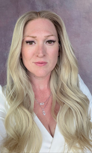 A woman with long blonde hair wearing a white top and a silver rectangular pendant necklace, posing in front of a textured mauve-purple backdrop.