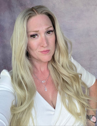 A blonde woman with long wavy hair wearing a white top and silver necklace, posing against a textured purple studio backdrop.