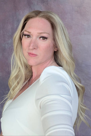 A blonde woman wearing a white long-sleeve top, photographed in a mirror selfie style against a textured purple backdrop.