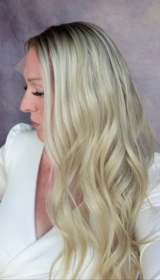 A woman with long, wavy platinum blonde hair is shown in profile against a textured mauve-purple studio backdrop, wearing a white top.