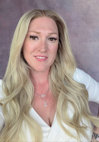 A woman with long blonde hair wearing a white top and a silver pendant necklace, posing in front of a textured mauve backdrop.